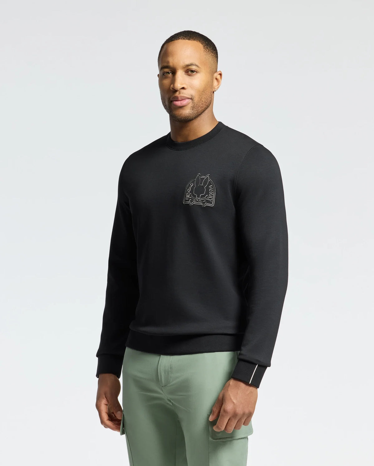 A person wearing a Psycho Bunny MENS MELBOURNE CLASSIC CREWNECK SWEATSHIRT - B6S561F200, featuring a subtle chest design, stands confidently. The soft stretch jersey and breathable Modal blend ensure comfort. They pair it with light green pants against a plain white background.