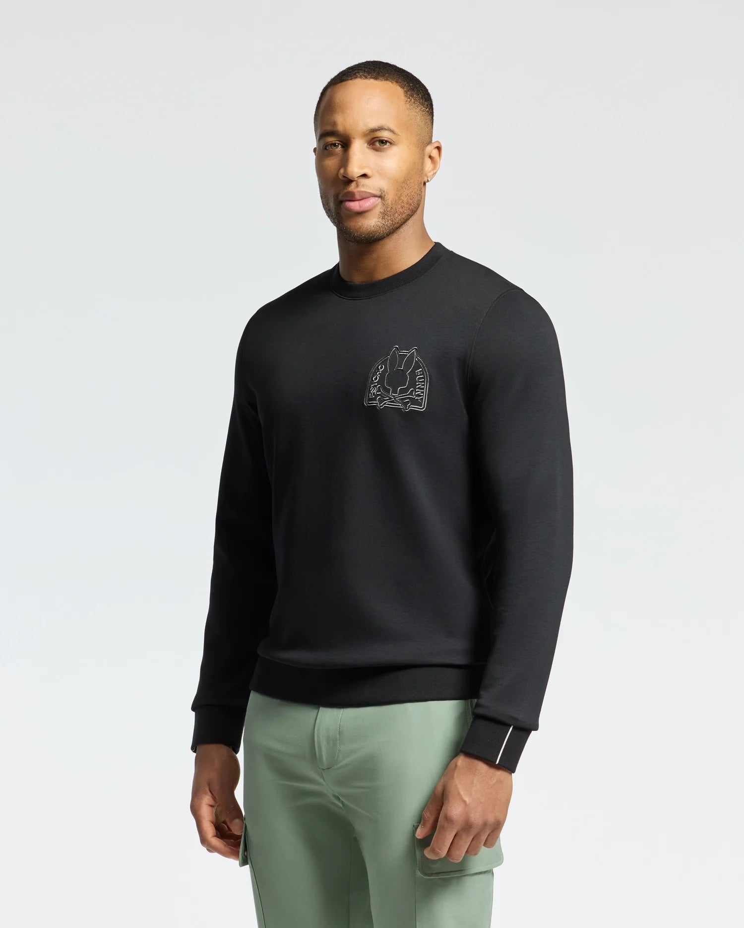 A person wearing a Psycho Bunny MENS MELBOURNE CLASSIC CREWNECK SWEATSHIRT - B6S561F200, featuring a subtle chest design, stands confidently. The soft stretch jersey and breathable Modal blend ensure comfort. They pair it with light green pants against a plain white background.
