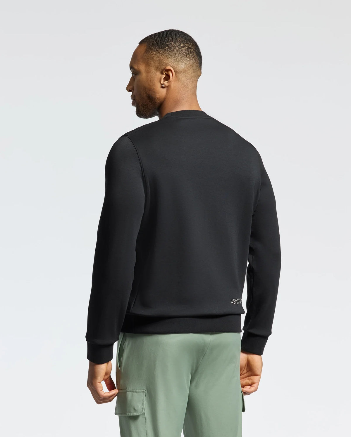 A man stands facing away from the camera in a Psycho Bunny MENS MELBOURNE CLASSIC CREWNECK SWEATSHIRT - B6S561F200, made from breathable Modal blend. Paired with green cargo pants, he contrasts perfectly against the light background, highlighting its effortless style.