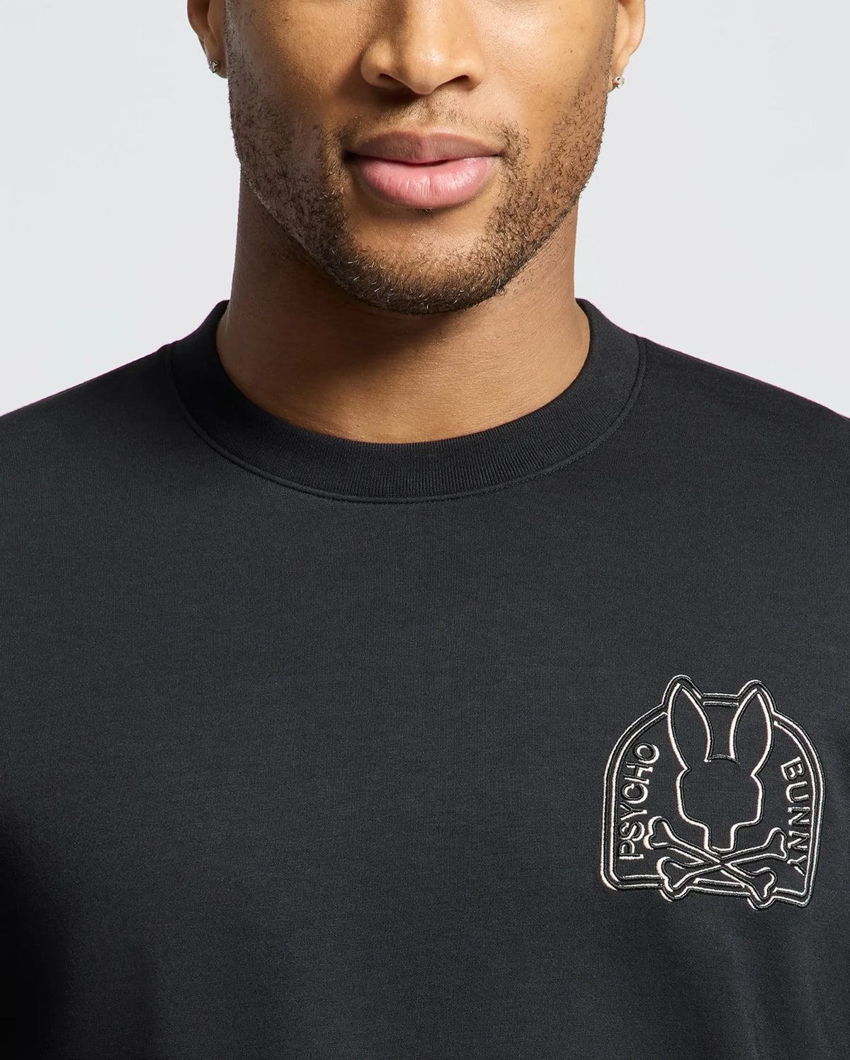 A man is wearing a Psycho Bunny Men's Melbourne Classic Crewneck Sweatshirt (B6S561F200) in black, made from a breathable Modal blend, featuring an embroidered rabbit and crossbones patch. Only his lower face and upper torso are visible against a plain white background.