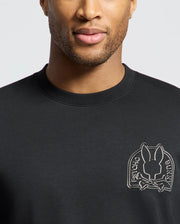 A man is wearing a Psycho Bunny Men's Melbourne Classic Crewneck Sweatshirt (B6S561F200) in black, made from a breathable Modal blend, featuring an embroidered rabbit and crossbones patch. Only his lower face and upper torso are visible against a plain white background.