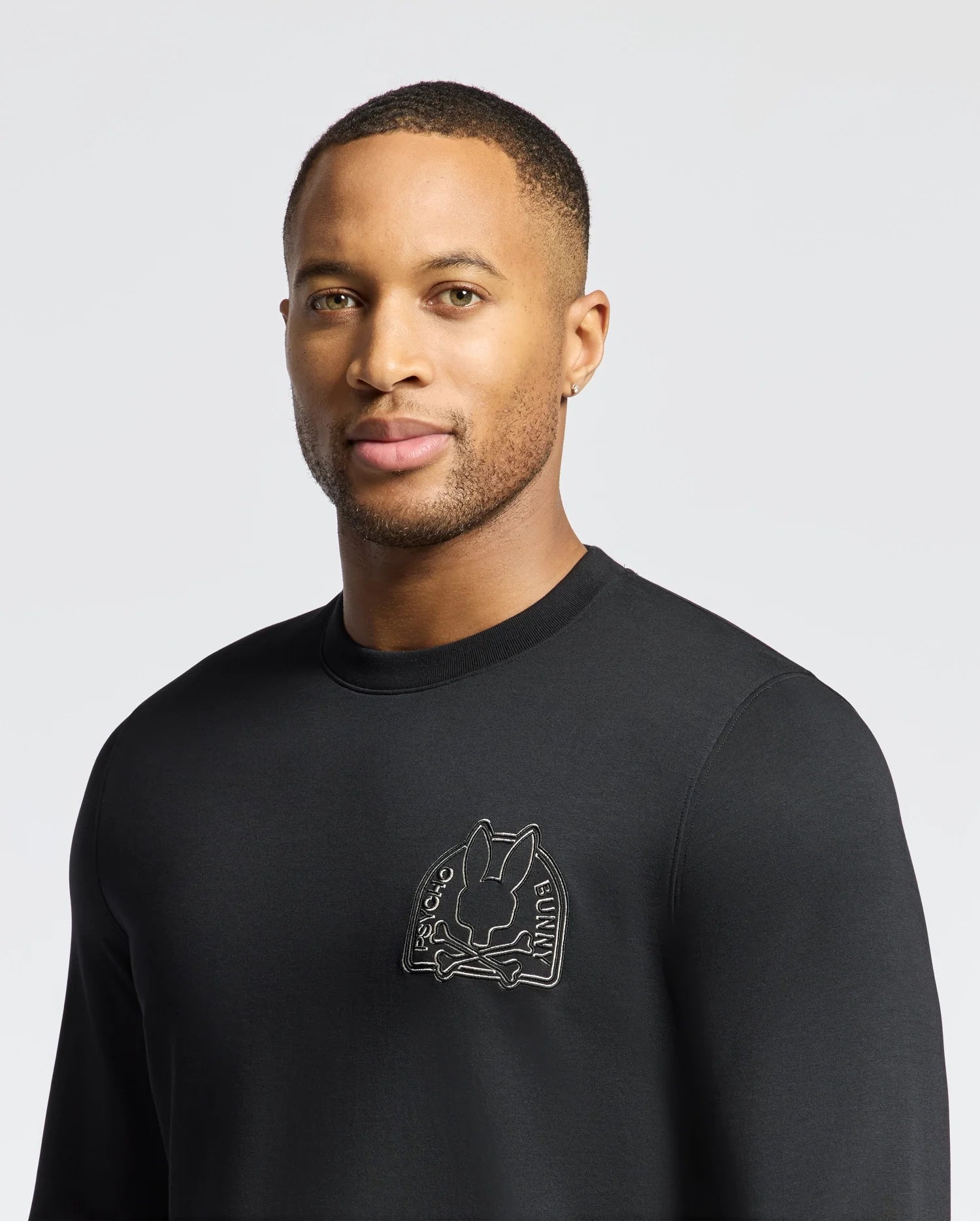 A person in a black Psycho Bunny Melbourne Classic Crewneck Sweatshirt featuring a rabbit graphic, stands against a gray background with short hair and a neutral expression, showcasing style and comfort in breathable Modal blend fabric.