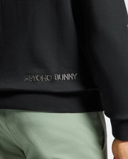 A close-up of a person in Psycho Bunny's black jacket features intricate white embroidery, perfectly paired with light green pants. The MENS MELBOURNE CLASSIC CREWNECK SWEATSHIRT in soft stretch jersey offers comfort and style, while the breathable Modal blend ensures all-day ease.