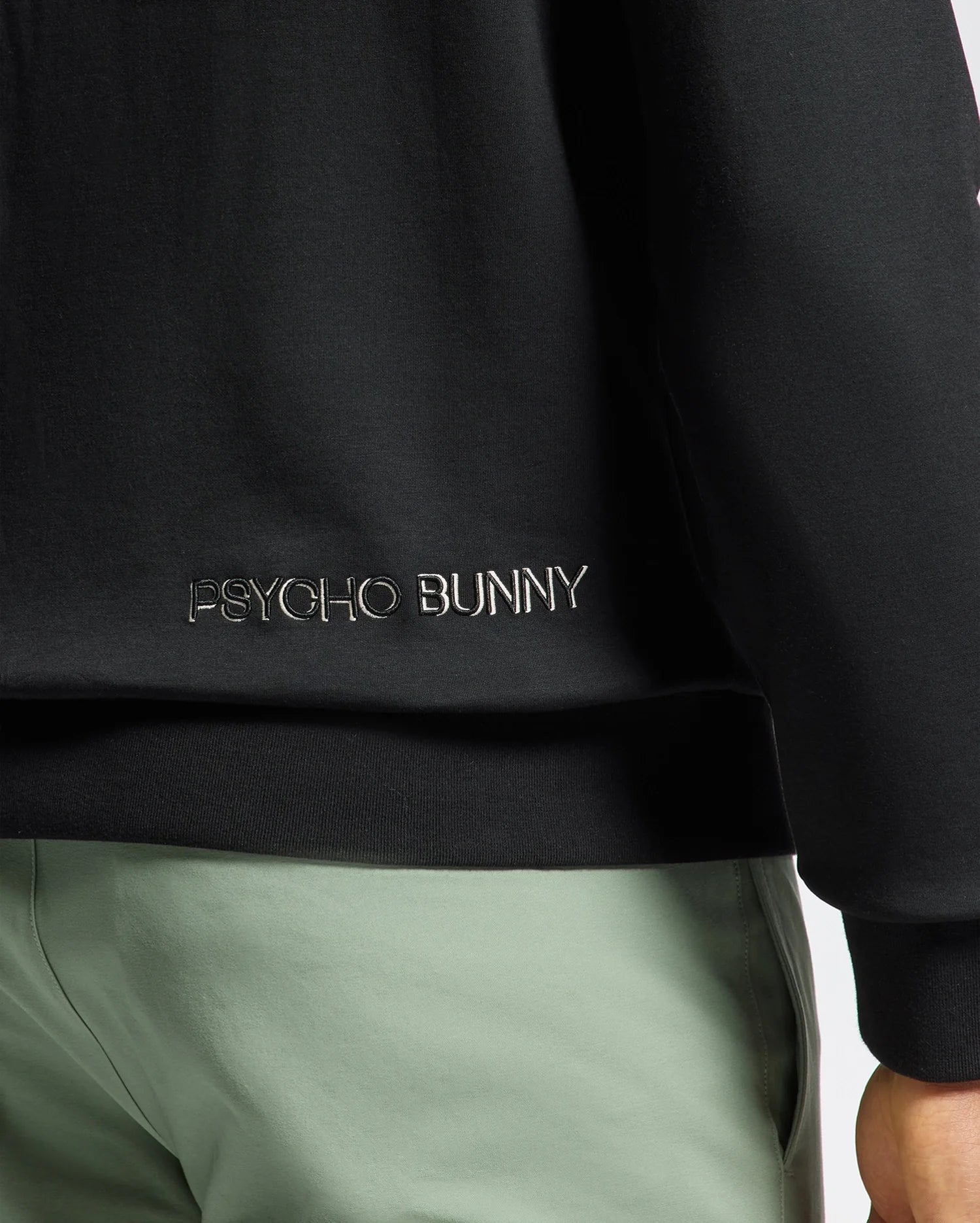 A close-up of a person in Psycho Bunny's black jacket features intricate white embroidery, perfectly paired with light green pants. The MENS MELBOURNE CLASSIC CREWNECK SWEATSHIRT in soft stretch jersey offers comfort and style, while the breathable Modal blend ensures all-day ease.