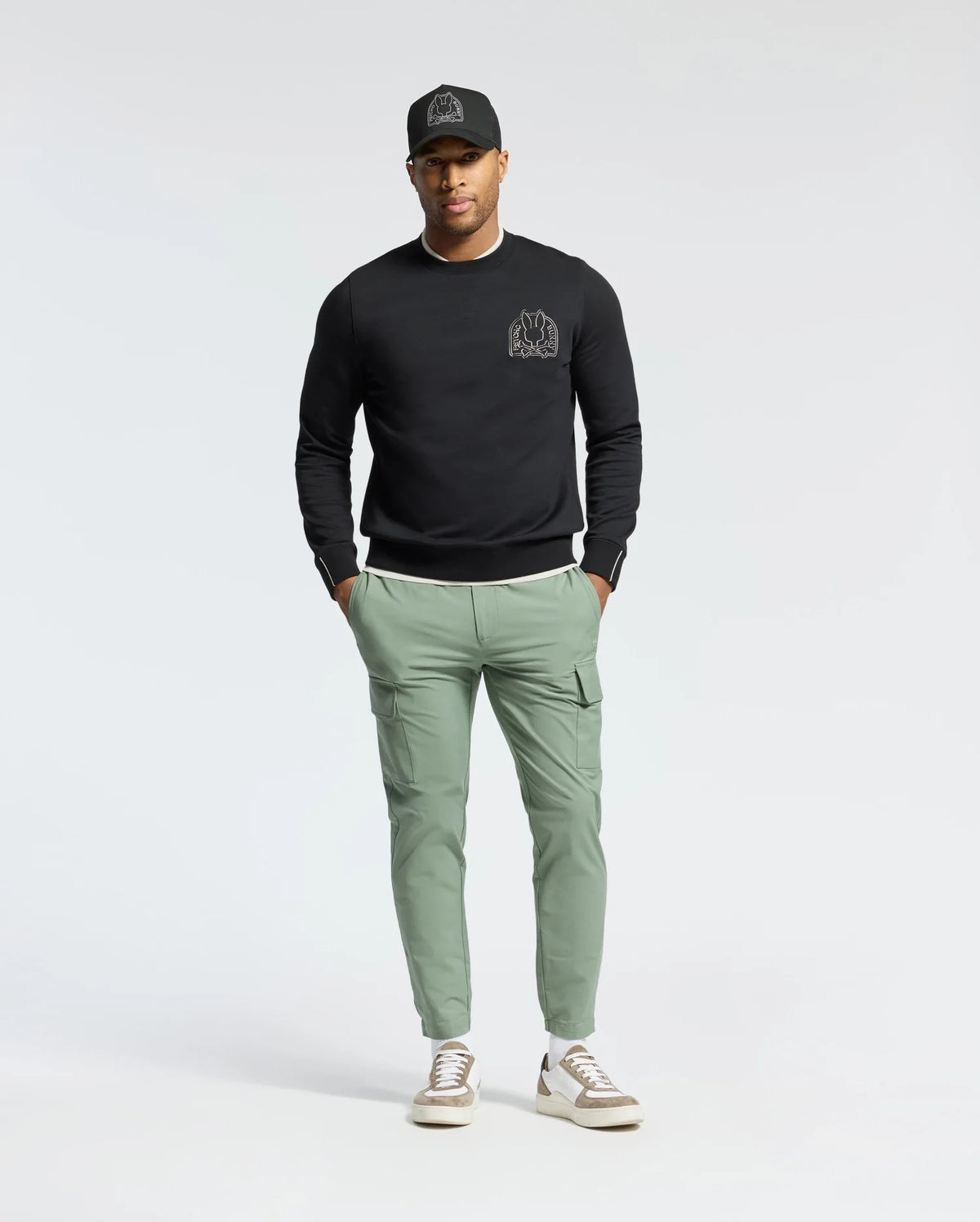 A person stands casually with hands in pockets against a plain background, wearing a black cap, Psycho Bunny MENS MELBOURNE CLASSIC CREWNECK SWEATSHIRT (B6S561F200) with a small chest design, light green cargo pants, and white sneakers.
