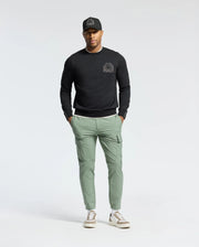 A person stands casually with hands in pockets against a plain background, wearing a black cap, Psycho Bunny MENS MELBOURNE CLASSIC CREWNECK SWEATSHIRT (B6S561F200) with a small chest design, light green cargo pants, and white sneakers.
