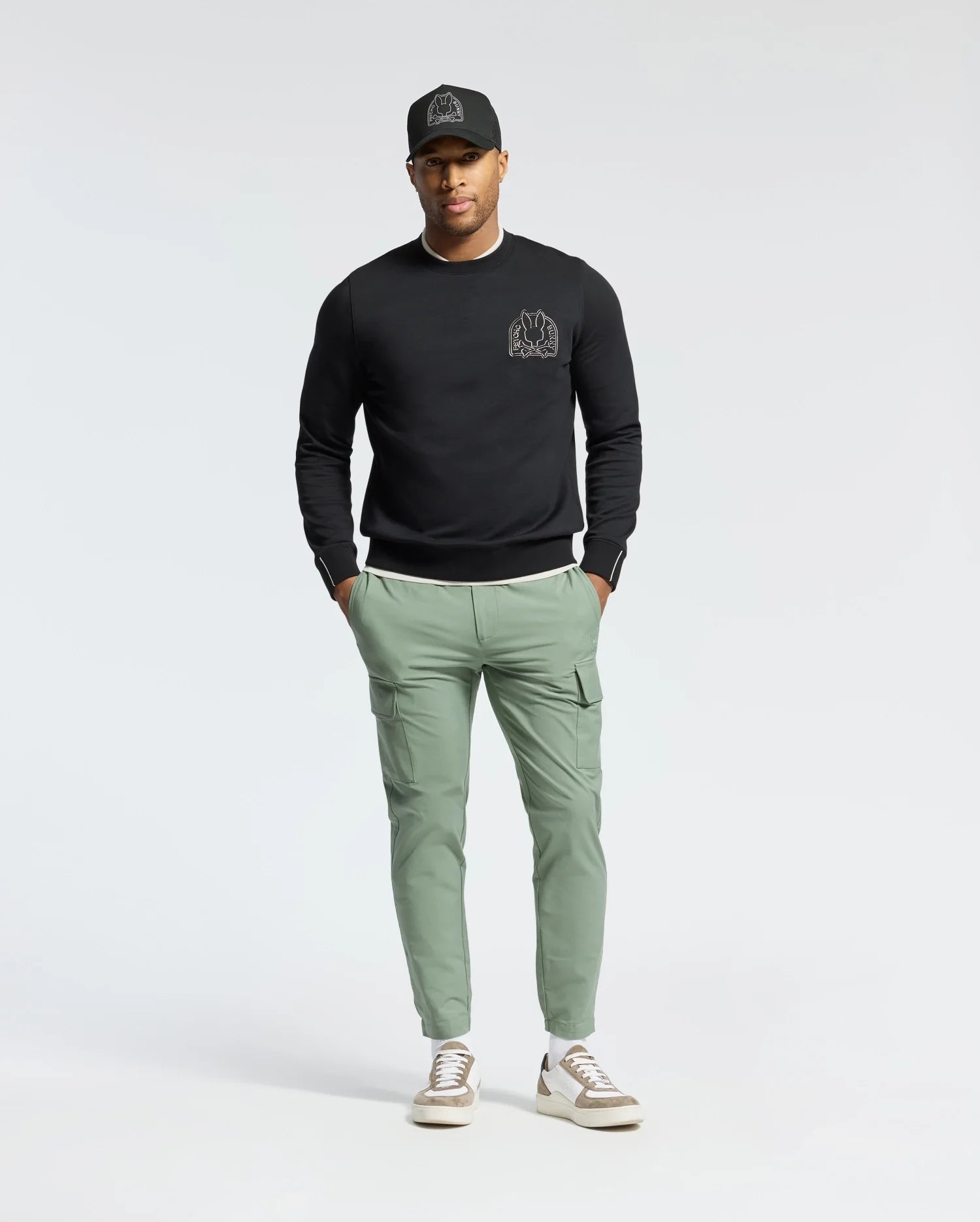 A person stands casually with hands in pockets against a plain background, wearing a black cap, Psycho Bunny MENS MELBOURNE CLASSIC CREWNECK SWEATSHIRT (B6S561F200) with a small chest design, light green cargo pants, and white sneakers.