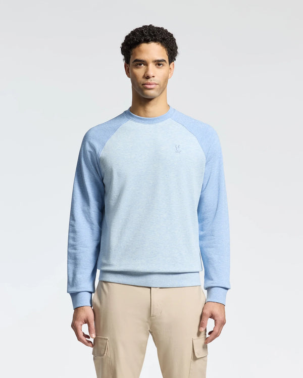 A person in a Psycho Bunny MENS ARCHIE SWEATSHIRT, B6S630F200, mixes light blue and navy tones with beige cargo pants, standing against a white backdrop for a monochrome look.