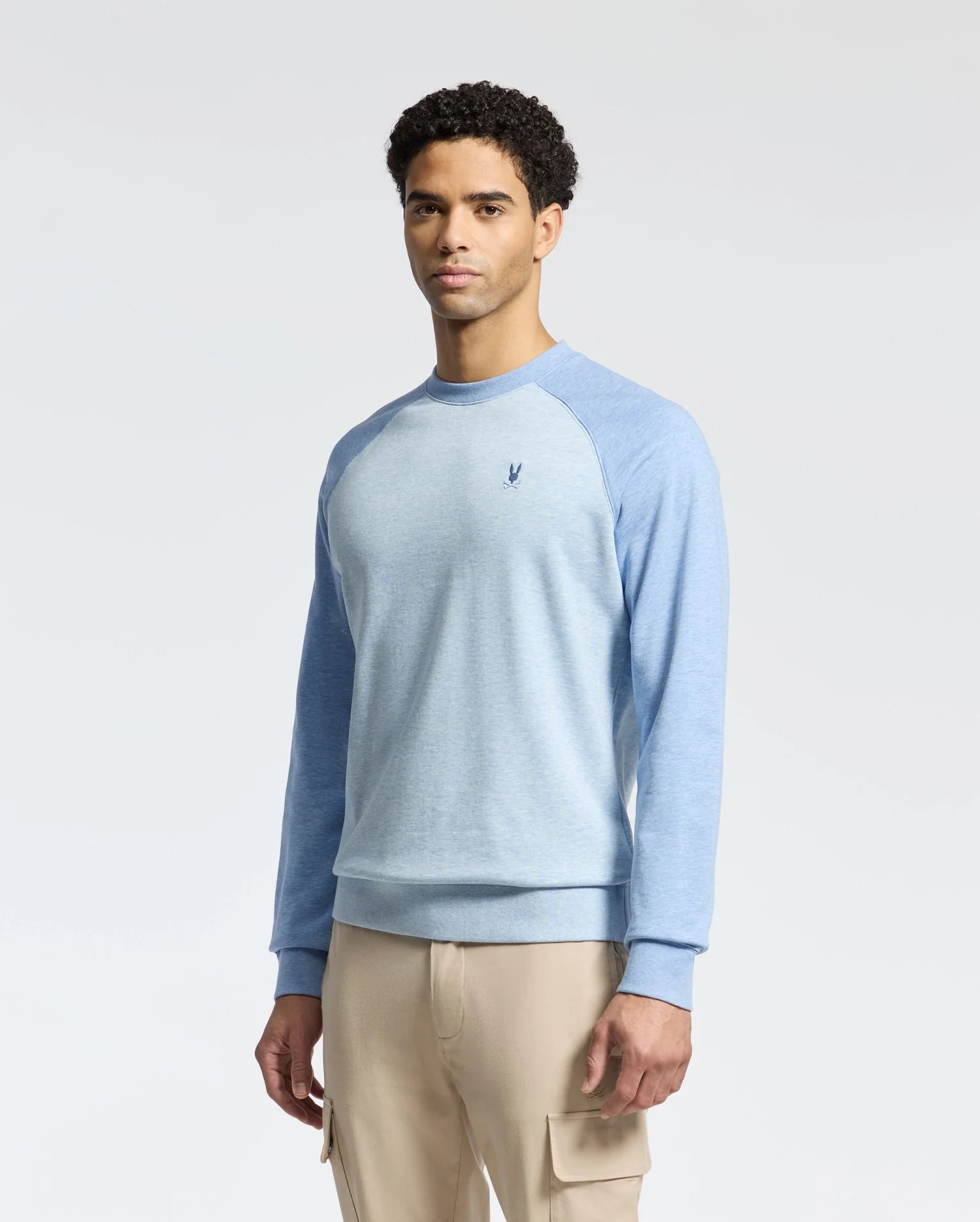 A person is wearing a Psycho Bunny MENS ARCHIE SWEATSHIRT - B6S630F200 in light blue with darker sleeves and beige cargo pants. The embroidered Bunny logo on the chest adds playfulness, while the plain white background enhances the monochrome style.