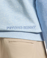 Close-up of a person in a light blue cotton MENS ARCHIE SWEATSHIRT - B6S630F200 by Psycho Bunny, with matching embroidery. They wear light tan pants, highlighting effortless monochrome styling.