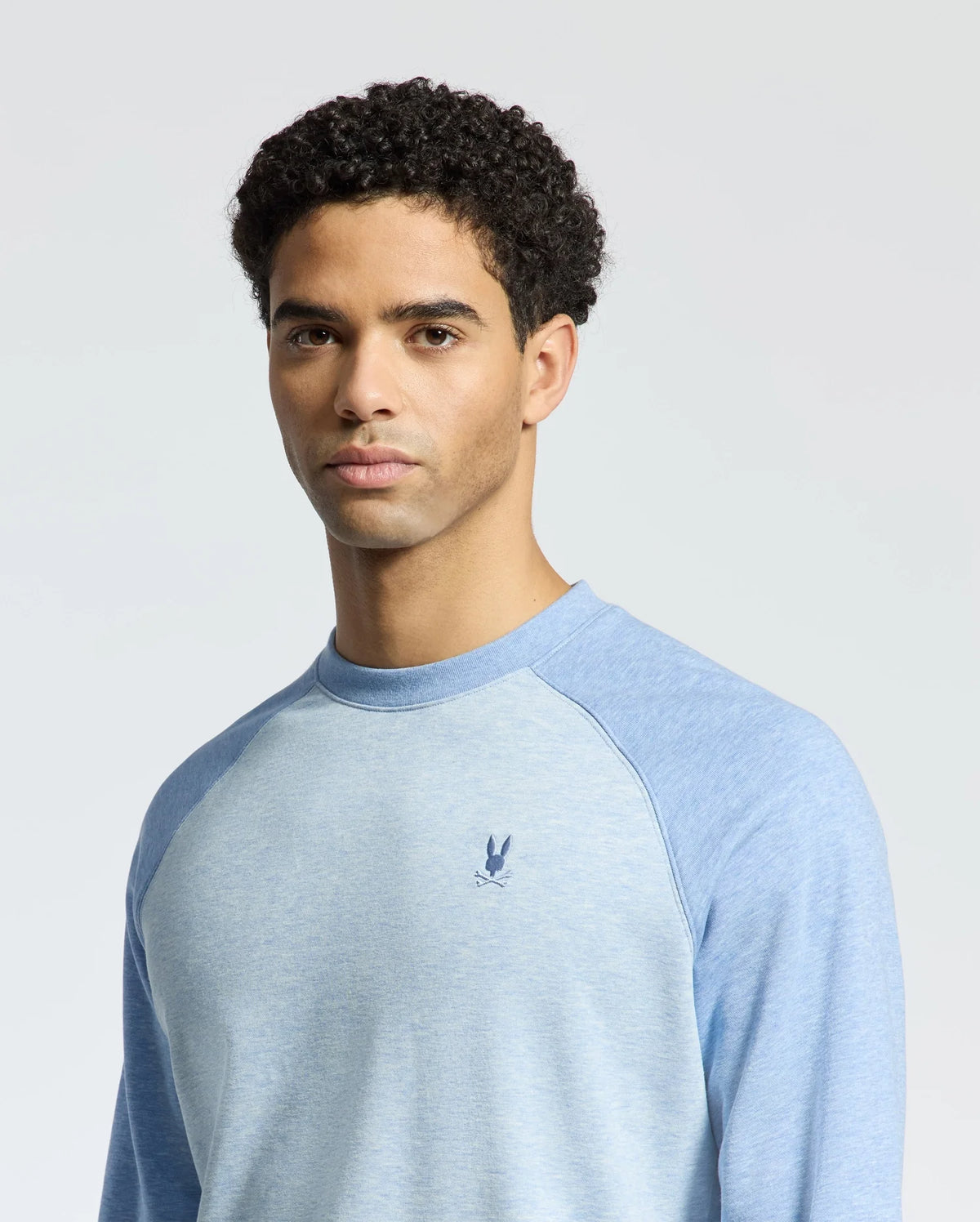 A person with short curly hair is wearing the Psycho Bunny MENS ARCHIE SWEATSHIRT - B6S630F200, a light blue cotton long-sleeved jersey featuring an embroidered Bunny logo on the chest. The plain light gray background enhances the monochrome styling.