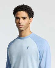 A person with short curly hair is wearing the Psycho Bunny MENS ARCHIE SWEATSHIRT - B6S630F200, a light blue cotton long-sleeved jersey featuring an embroidered Bunny logo on the chest. The plain light gray background enhances the monochrome styling.