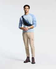 A man in a light blue Psycho Bunny MENS ARCHIE SWEATSHIRT - B6S630F200 and beige cargo pants stands against a plain background. He carries a black crossbody bag, wears brown boots and a watch, with his short, curly hair completing the monochrome look.
