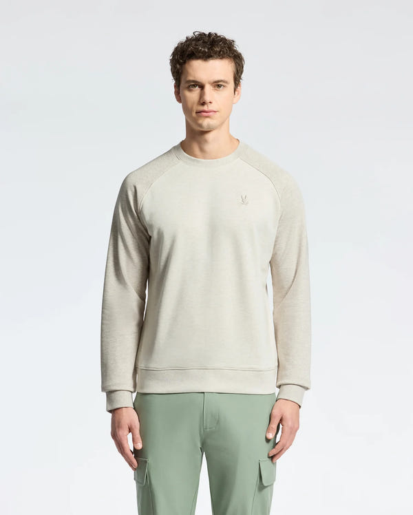A curly-haired man in Psycho Bunny's MENS ARCHIE MELANGE COLOR BLOCK SWEATSHIRT stands against a white background, pairing the light gray top with green cargo pants. The embroidered bunny on the left adds playful elegance.