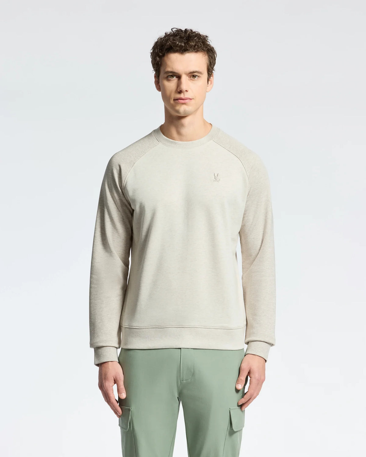 A curly-haired man in Psycho Bunny's MENS ARCHIE MELANGE COLOR BLOCK SWEATSHIRT stands against a white background, pairing the light gray top with green cargo pants. The embroidered bunny on the left adds playful elegance.