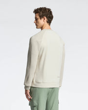 A person with curly hair wears a light beige Psycho Bunny Archie Melange Color Block sweatshirt and green cargo pants, standing against a plain background, gazing to the side.
