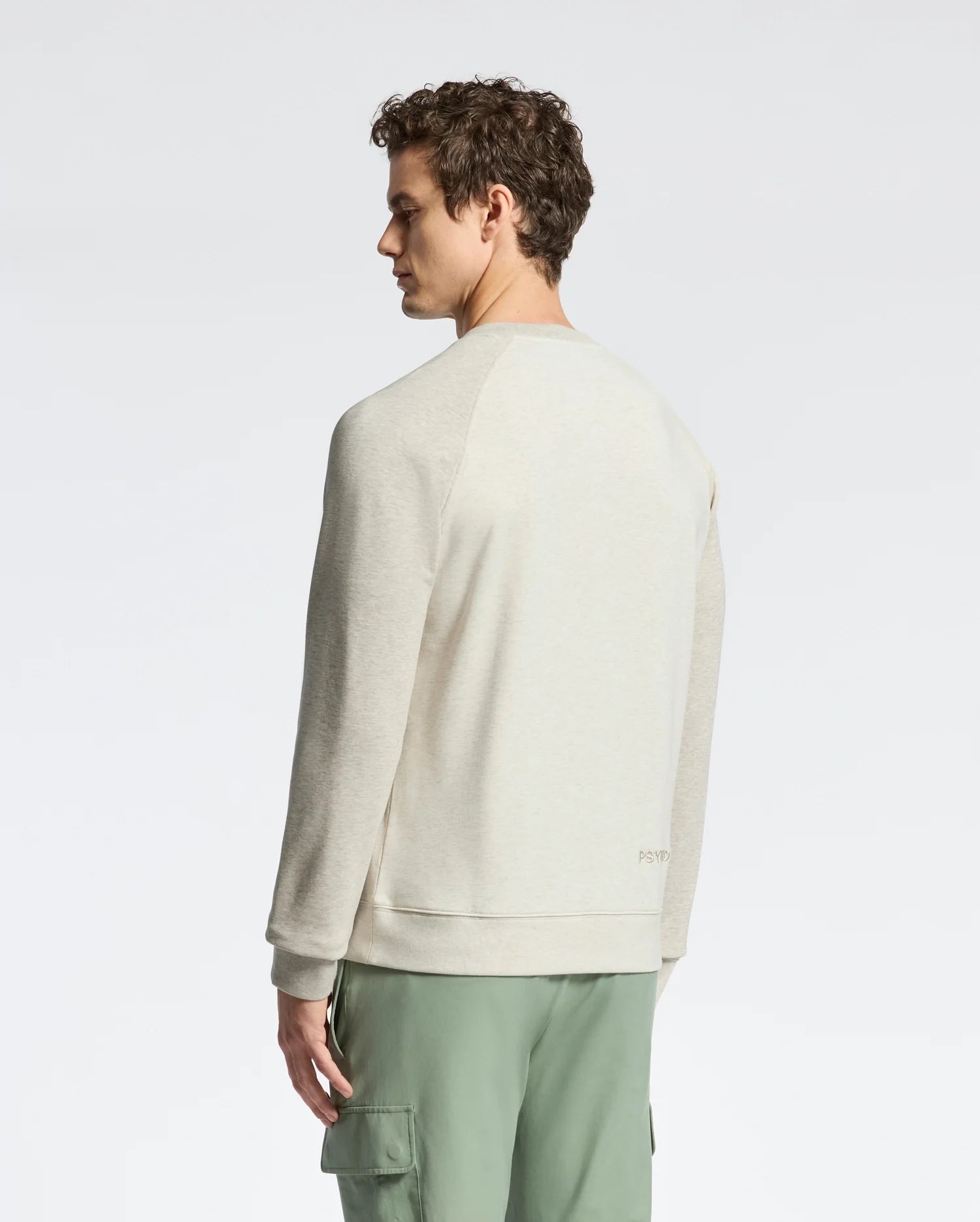 A person with curly hair wears a light beige Psycho Bunny Archie Melange Color Block sweatshirt and green cargo pants, standing against a plain background, gazing to the side.