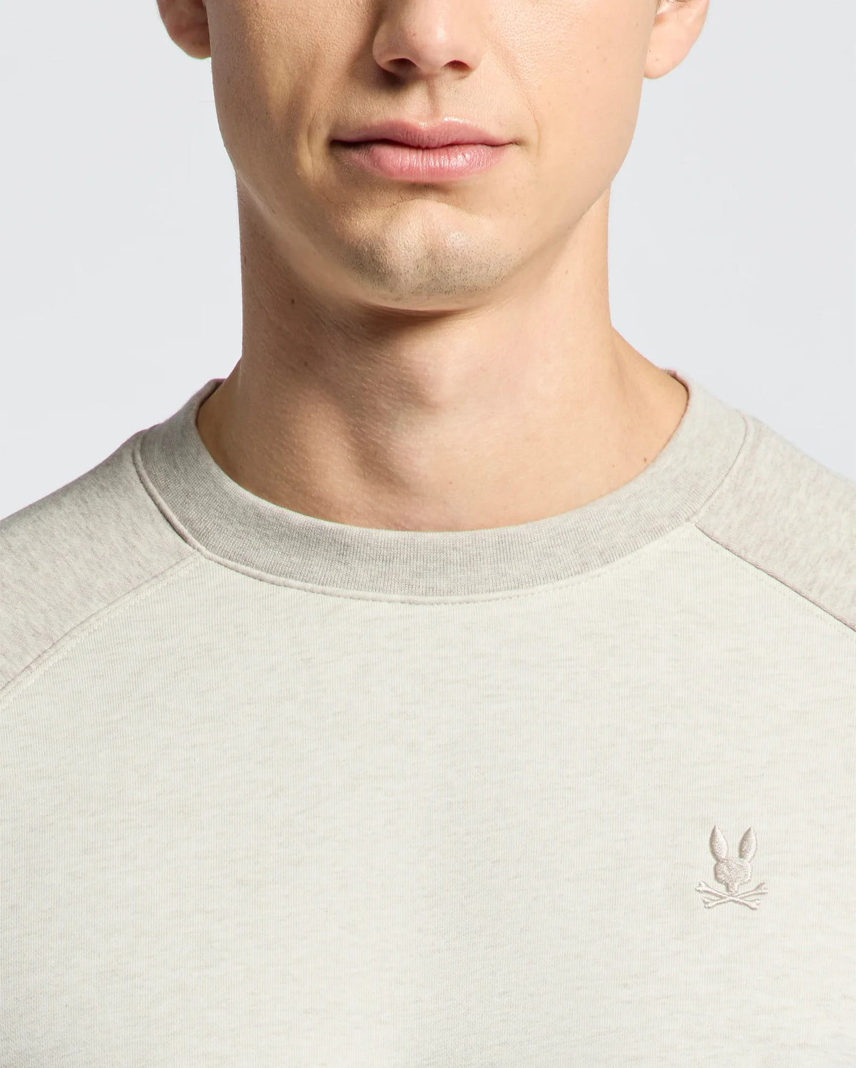 A person wearing the Psycho Bunny MENS ARCHIE MELANGE COLOR BLOCK SWEATSHIRT - B6S630F200 in light grey with a Bunny emblem embroidered on the left chest. The photo is cropped to focus on the lower face and upper chest, showcasing a monochrome style.