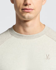 A person wearing the Psycho Bunny MENS ARCHIE MELANGE COLOR BLOCK SWEATSHIRT - B6S630F200 in light grey with a Bunny emblem embroidered on the left chest. The photo is cropped to focus on the lower face and upper chest, showcasing a monochrome style.