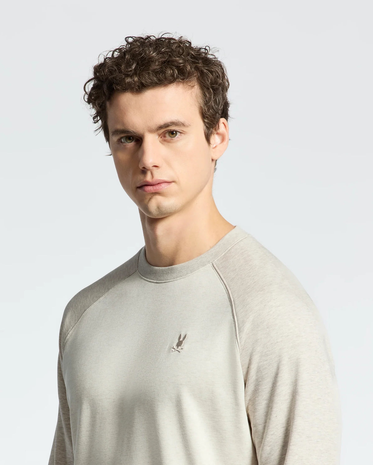 A curly-haired young man models a Psycho Bunny MENS ARCHIE MELANGE COLOR BLOCK SWEATSHIRT, B6S630F200, against a plain background for monochrome styling.