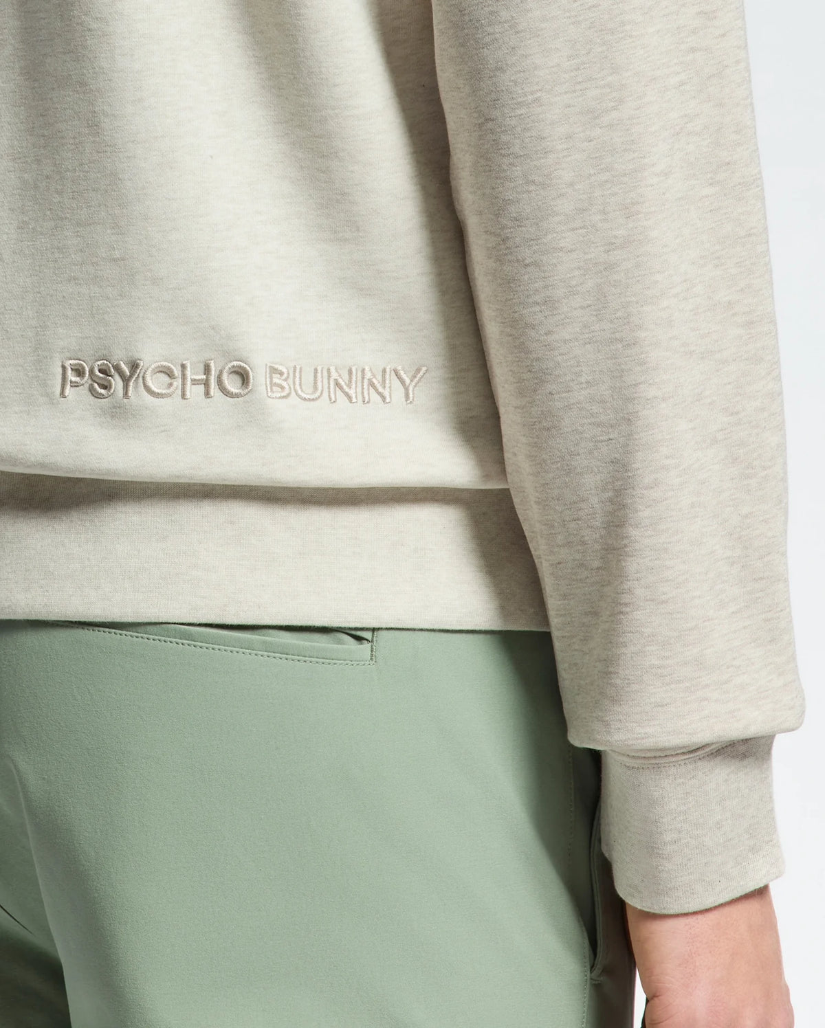 Close-up of a person wearing the Psycho Bunny MENS ARCHIE MELANGE COLOR BLOCK SWEATSHIRT - B6S630F200 in light beige, with "PSYCHO BUNNY" and a Bunny design embroidered. It's paired with light green pants for a subtle color-blocked look, focusing on the lower back and arm area.