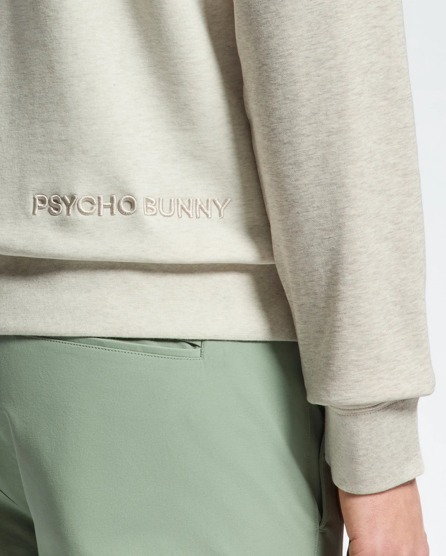 Close-up of a person wearing the Psycho Bunny MENS ARCHIE MELANGE COLOR BLOCK SWEATSHIRT - B6S630F200 in light beige, with "PSYCHO BUNNY" and a Bunny design embroidered. It's paired with light green pants for a subtle color-blocked look, focusing on the lower back and arm area.