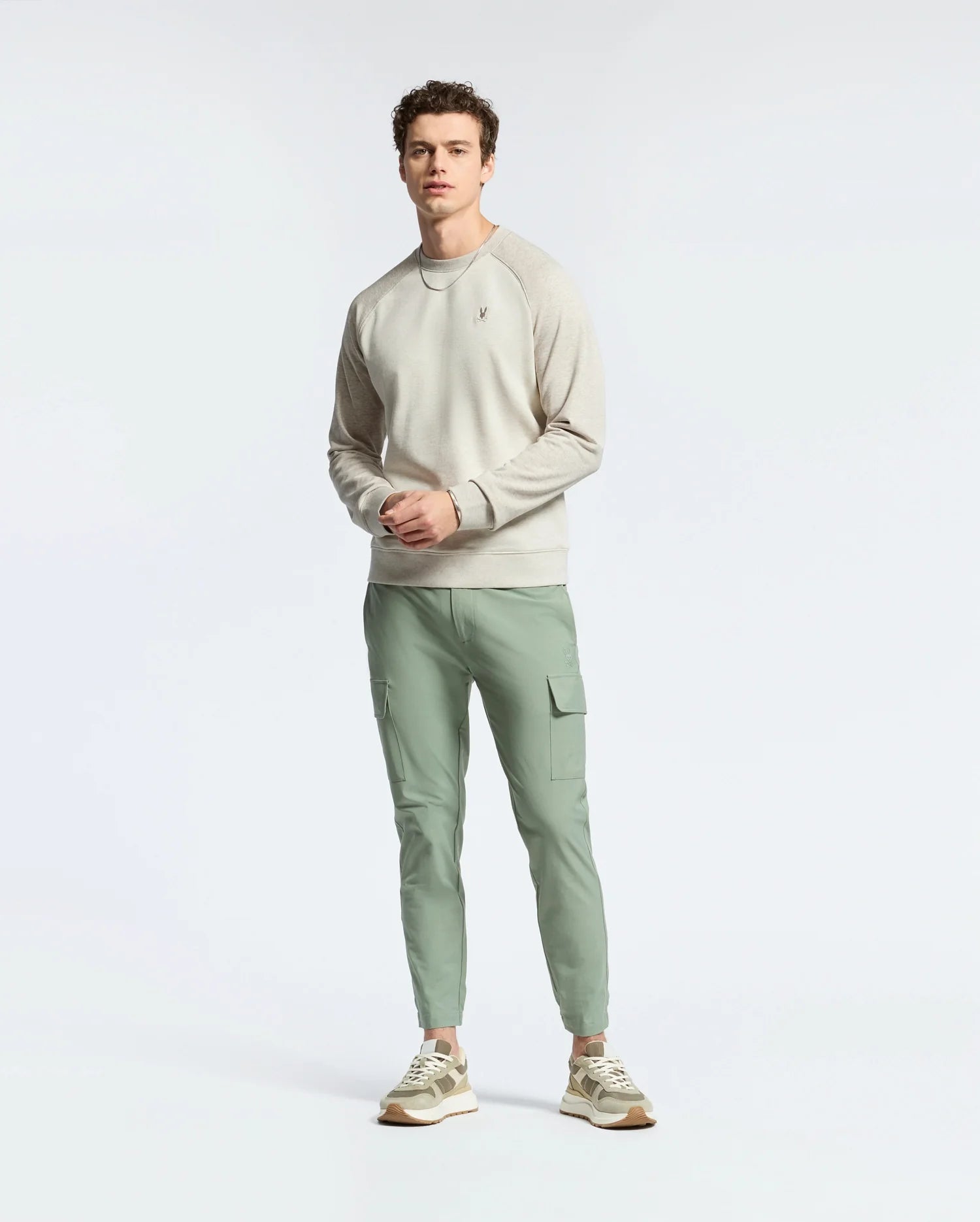 A person stands against a light background in the Psycho Bunny MENS ARCHIE MELANGE COLOR BLOCK SWEATSHIRT - B6S630F200, light green cargo pants, and sneakers. They have short, curly hair and a neutral expression while looking at the camera.