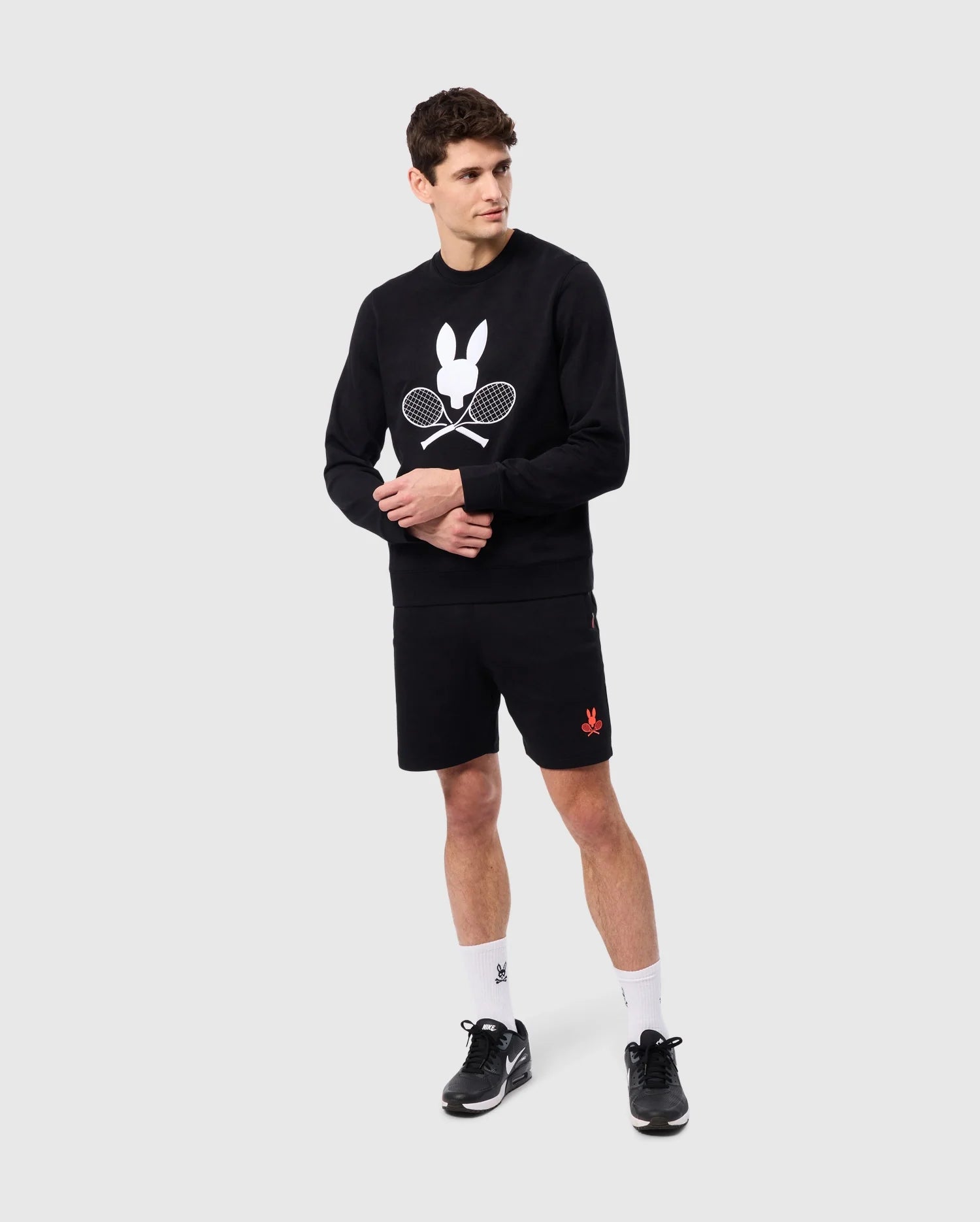 A man stands against a plain background wearing the tennis-inspired MENS COURTSIDE SWEATSHIRT - B6S688C200 by Psycho Bunny, featuring a rabbit and racquet design. He pairs it with black shorts, white socks, and sleek sneakers. Made from a breathable cotton-blend fabric, he clasps his hands while gazing thoughtfully to the side.
