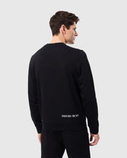 An individual dressed in a black MENS COURTSIDE SWEATSHIRT - B6S688C200 and matching pants faces away from the camera. The breathable cotton-blend sweatshirt boasts the "Psycho Bunny" branding on the lower back. The plain light gray backdrop enhances the minimalist style of this tennis-inspired ensemble.