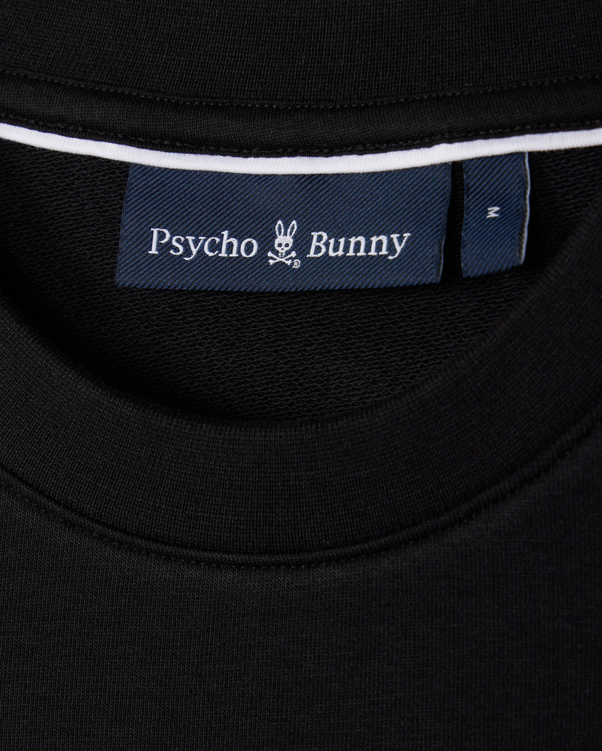 A close-up of the black Men's Courtside Sweatshirt's neckline showcasing a blue label with a white bunny logo and "Psycho Bunny" branding. Made from a breathable cotton-blend, this piece fuses courtside sweatshirt style with tennis-inspired flair.