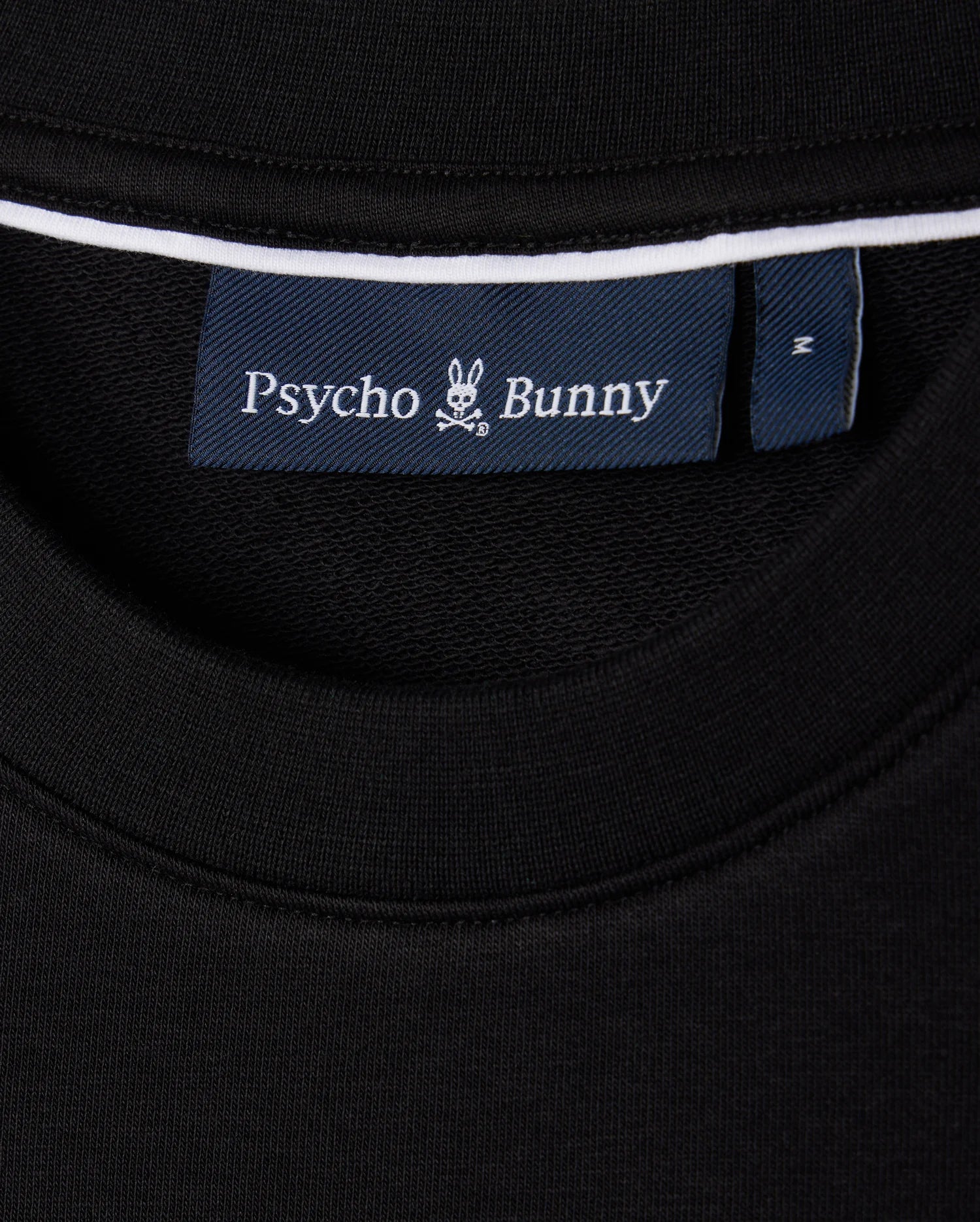 A close-up of the black Men's Courtside Sweatshirt's neckline showcasing a blue label with a white bunny logo and "Psycho Bunny" branding. Made from a breathable cotton-blend, this piece fuses courtside sweatshirt style with tennis-inspired flair.