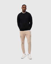 A man stands against a gray background, wearing the stylish MENS DUKE EMBROIDERED SWEATSHIRT by Psycho Bunny, paired with beige cargo pants and white sneakers. With his hands in his pockets, he exudes a relaxed vibe.