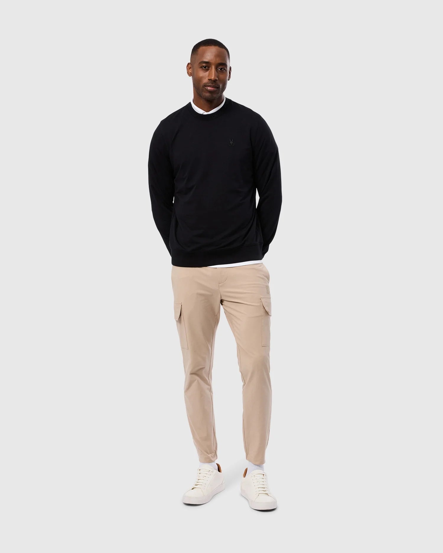 A man stands against a gray background, wearing the stylish MENS DUKE EMBROIDERED SWEATSHIRT by Psycho Bunny, paired with beige cargo pants and white sneakers. With his hands in his pockets, he exudes a relaxed vibe.