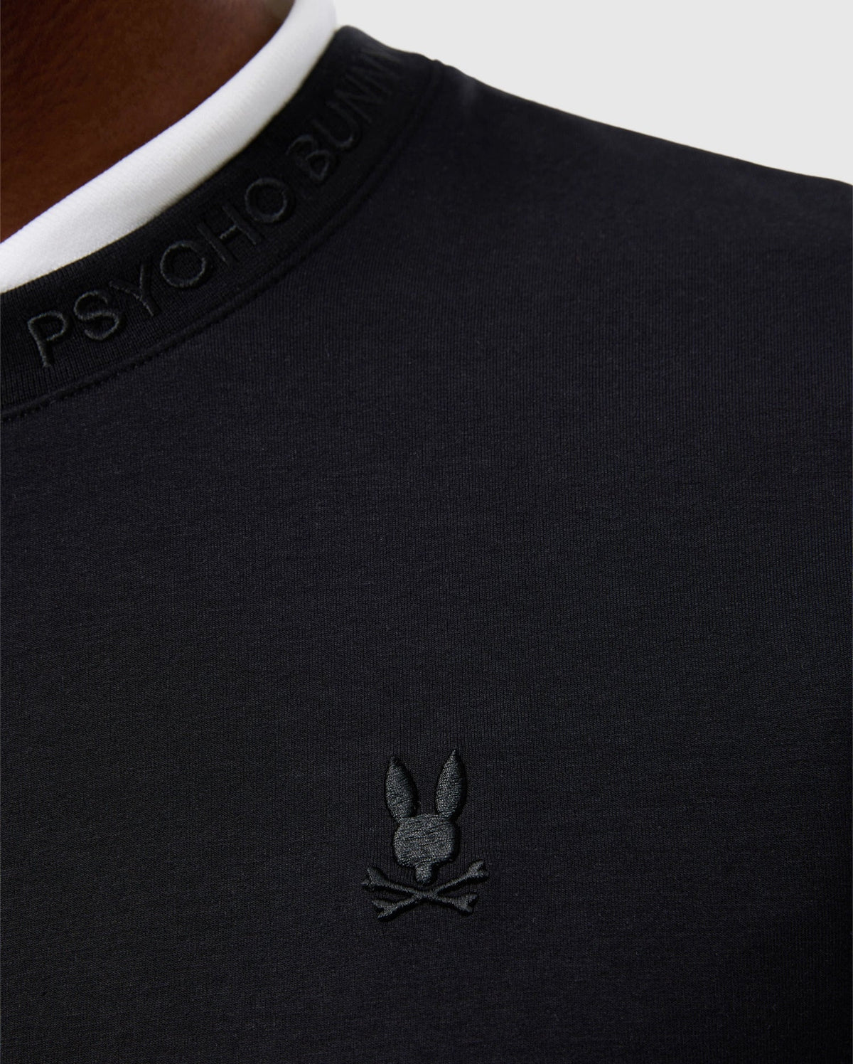 Close-up of a person wearing the black cotton-blend MENS DUKE EMBROIDERED SWEATSHIRT - B6S706D200 by Psycho Bunny, featuring a white collar. This luxury pullover showcases the brand's logo, a stylized bunny with crossbones, embroidered on the chest.