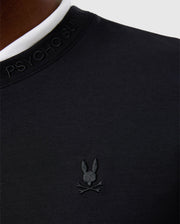 Close-up of a person wearing the black cotton-blend MENS DUKE EMBROIDERED SWEATSHIRT - B6S706D200 by Psycho Bunny, featuring a white collar. This luxury pullover showcases the brand's logo, a stylized bunny with crossbones, embroidered on the chest.