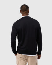 A man wearing a luxurious Psycho Bunny MENS DUKE EMBROIDERED SWEATSHIRT (B6S706D200) and beige cargo pants stands with his back to the camera against a light gray background.