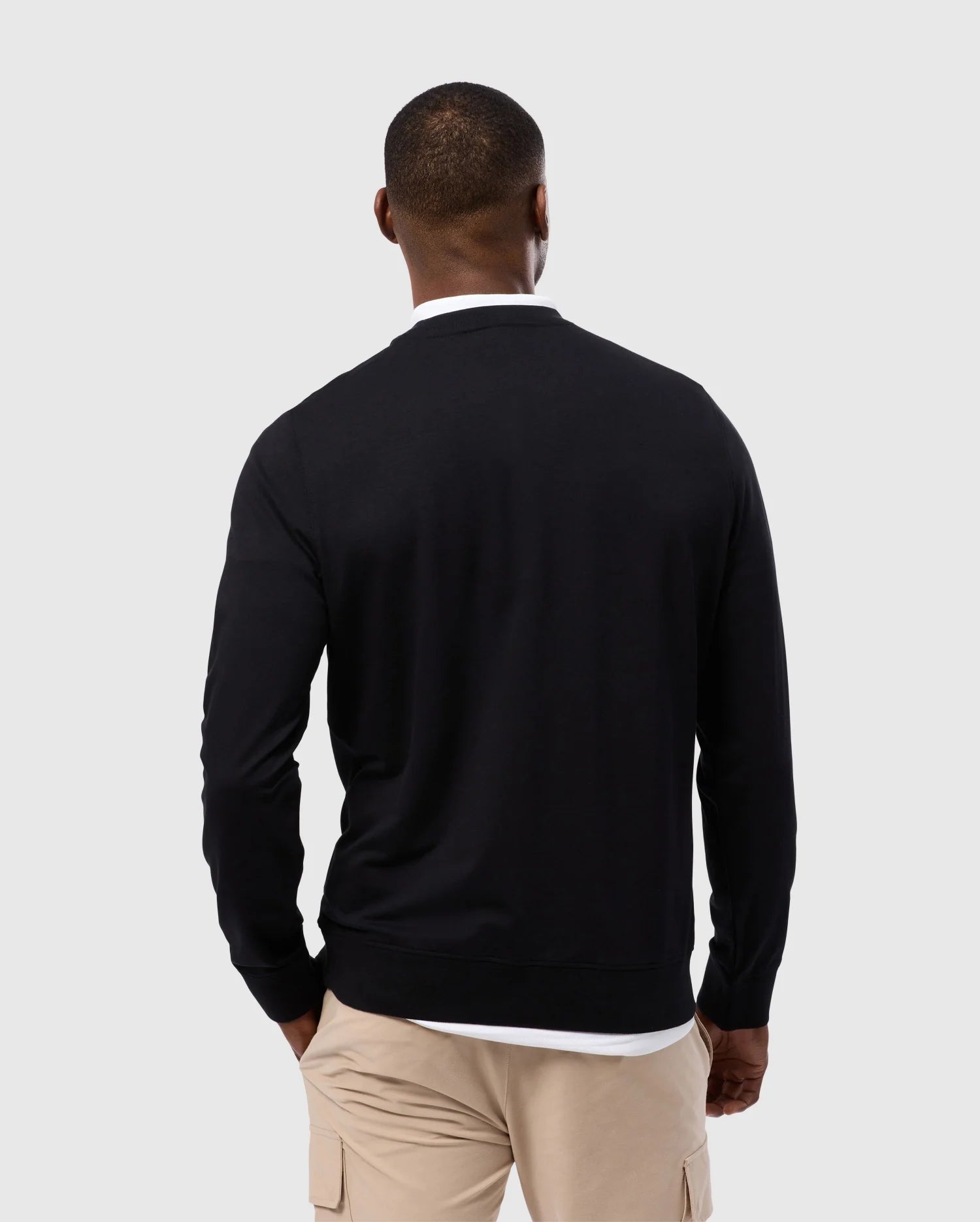 A man wearing a luxurious Psycho Bunny MENS DUKE EMBROIDERED SWEATSHIRT (B6S706D200) and beige cargo pants stands with his back to the camera against a light gray background.