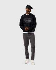 A man wearing the MENS TACOMA MODAL SWEATSHIRT by Psycho Bunny, characterized by its premium modal fabric, along with a black cap, gray pants, and white sneakers, stands against a plain background, smiling with his hands together.