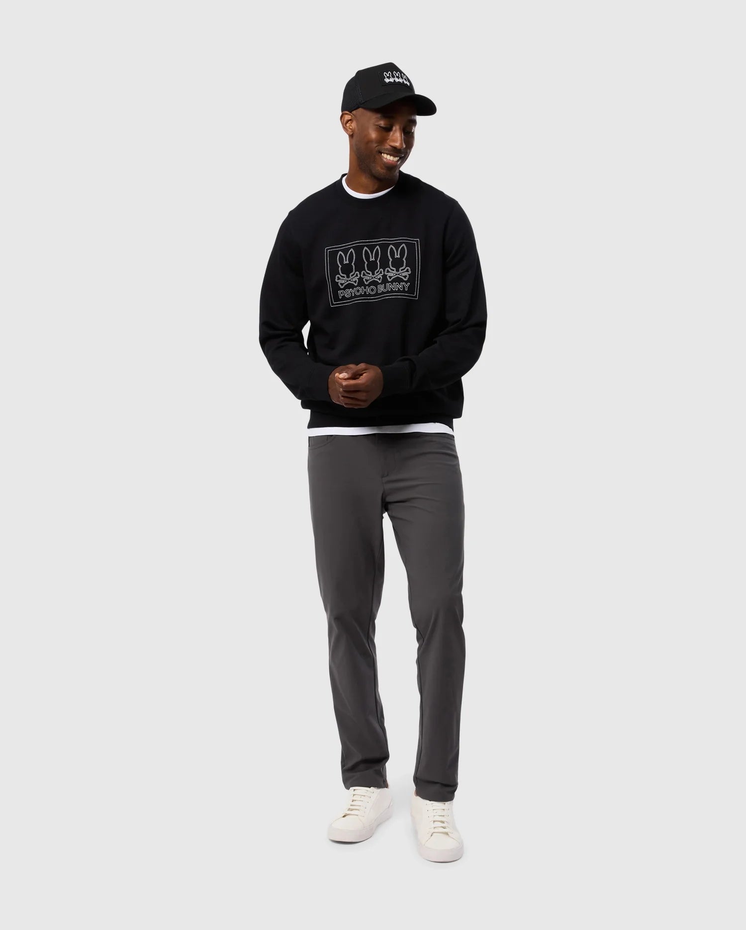 A man wearing the MENS TACOMA MODAL SWEATSHIRT by Psycho Bunny, characterized by its premium modal fabric, along with a black cap, gray pants, and white sneakers, stands against a plain background, smiling with his hands together.