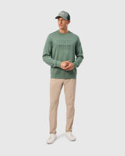A man wearing the Psycho Bunny MENS TACOMA SWEATSHIRT - B6S927D200 in green, along with beige trousers, white sneakers, and a coordinating green cap stands against a plain backdrop. His outfit, crafted from premium stretch fabric, promises luxurious comfort as he clasps his hands together in front of him.