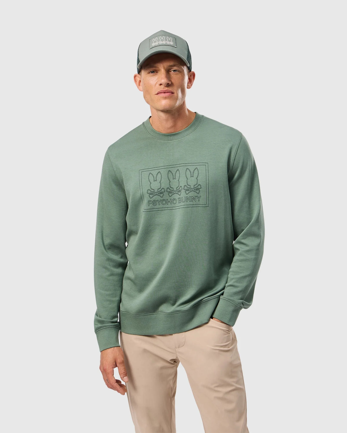 Dressed in the MENS TACOMA SWEATSHIRT by Psycho Bunny, a person stands against a plain white background. The green sweatshirt features a graphic of three bunnies and "PRAYING BUNNY" text. With their hand in the pocket of beige pants, they exemplify luxurious comfort and style, accessorized with a matching green cap.