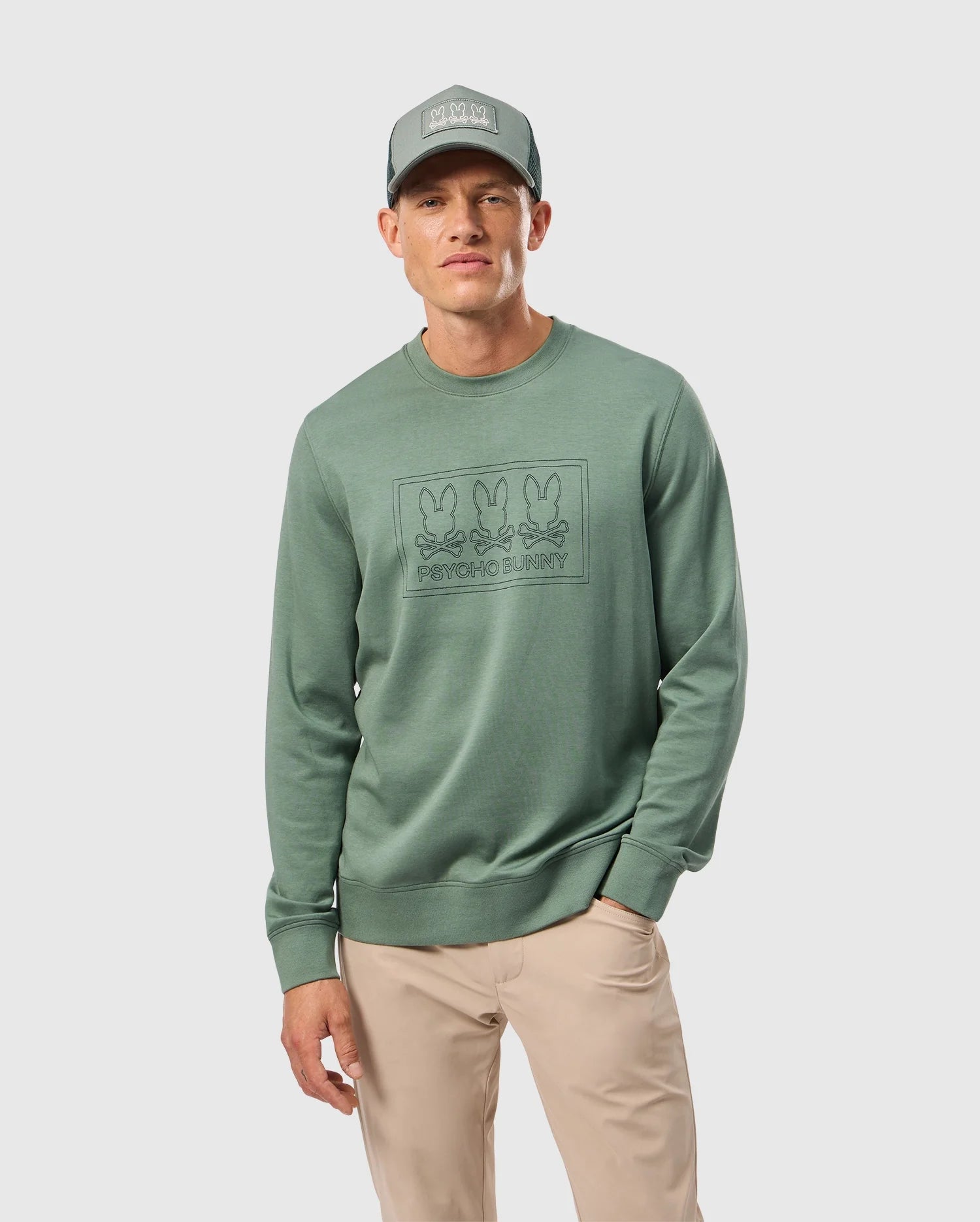 Dressed in the MENS TACOMA SWEATSHIRT by Psycho Bunny, a person stands against a plain white background. The green sweatshirt features a graphic of three bunnies and "PRAYING BUNNY" text. With their hand in the pocket of beige pants, they exemplify luxurious comfort and style, accessorized with a matching green cap.