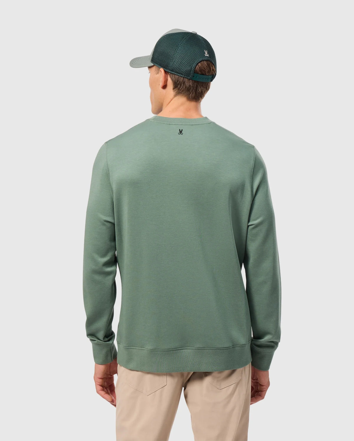 The image captures someone from behind wearing a Psycho Bunny MENS TACOMA SWEATSHIRT - B6S927D200 in green, embodying luxurious comfort with its premium stretch fabric. They complement the attire with beige trousers, set against a light gray background for a simple and neutral ambiance.