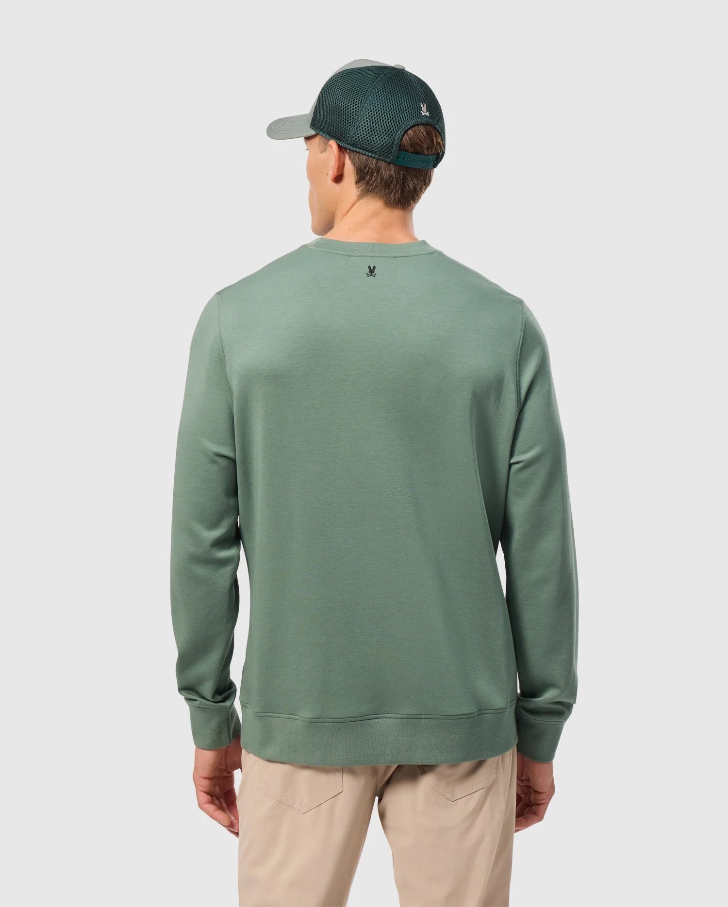 The image captures someone from behind wearing a Psycho Bunny MENS TACOMA SWEATSHIRT - B6S927D200 in green, embodying luxurious comfort with its premium stretch fabric. They complement the attire with beige trousers, set against a light gray background for a simple and neutral ambiance.