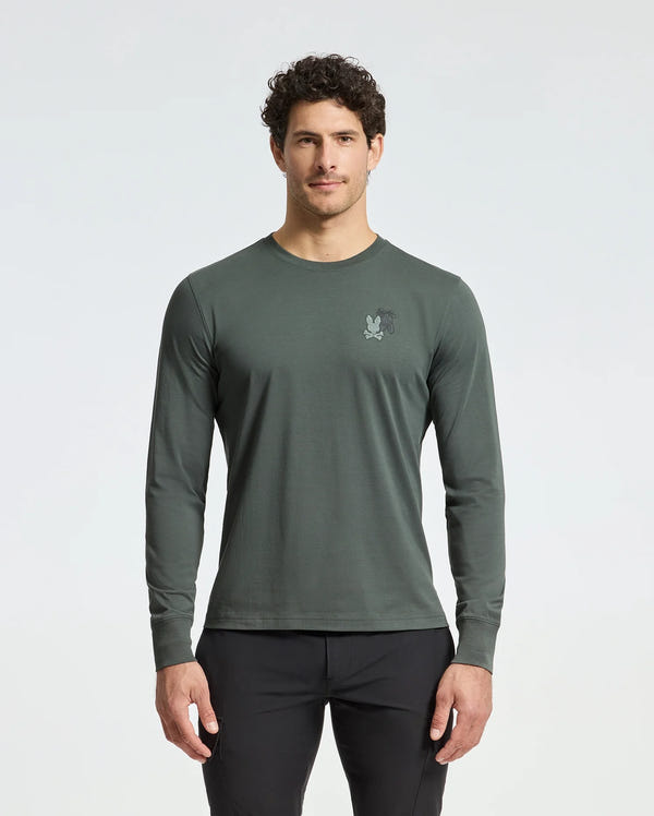 A man with short curly hair, wearing a Psycho Bunny dark green MENS PIERCE LONG SLEEVE BACK GRAPHIC TEE - B6T178E200 made from soft Pima cotton and featuring a small embroidered back graphic, stands against a light background. He's also wearing black pants.