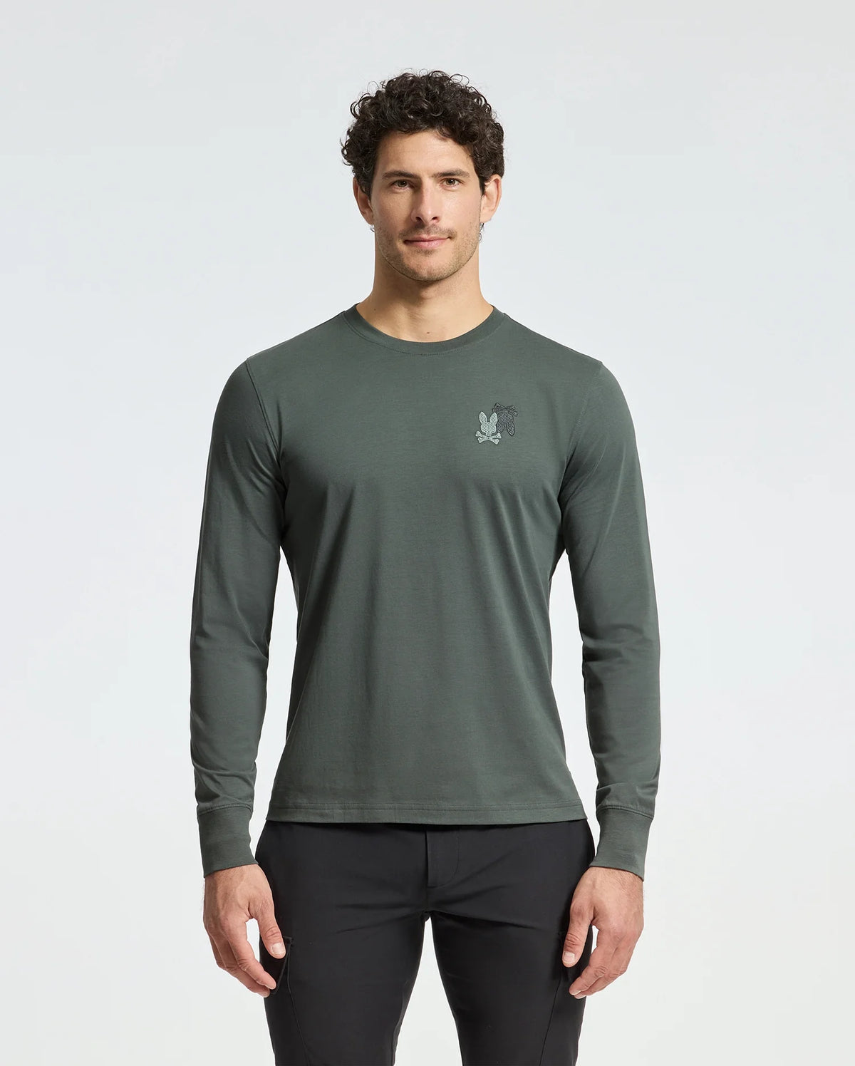 A man with short curly hair, wearing a Psycho Bunny dark green MENS PIERCE LONG SLEEVE BACK GRAPHIC TEE - B6T178E200 made from soft Pima cotton and featuring a small embroidered back graphic, stands against a light background. He's also wearing black pants.