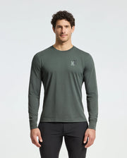 A man with short curly hair, wearing a Psycho Bunny dark green MENS PIERCE LONG SLEEVE BACK GRAPHIC TEE - B6T178E200 made from soft Pima cotton and featuring a small embroidered back graphic, stands against a light background. He's also wearing black pants.