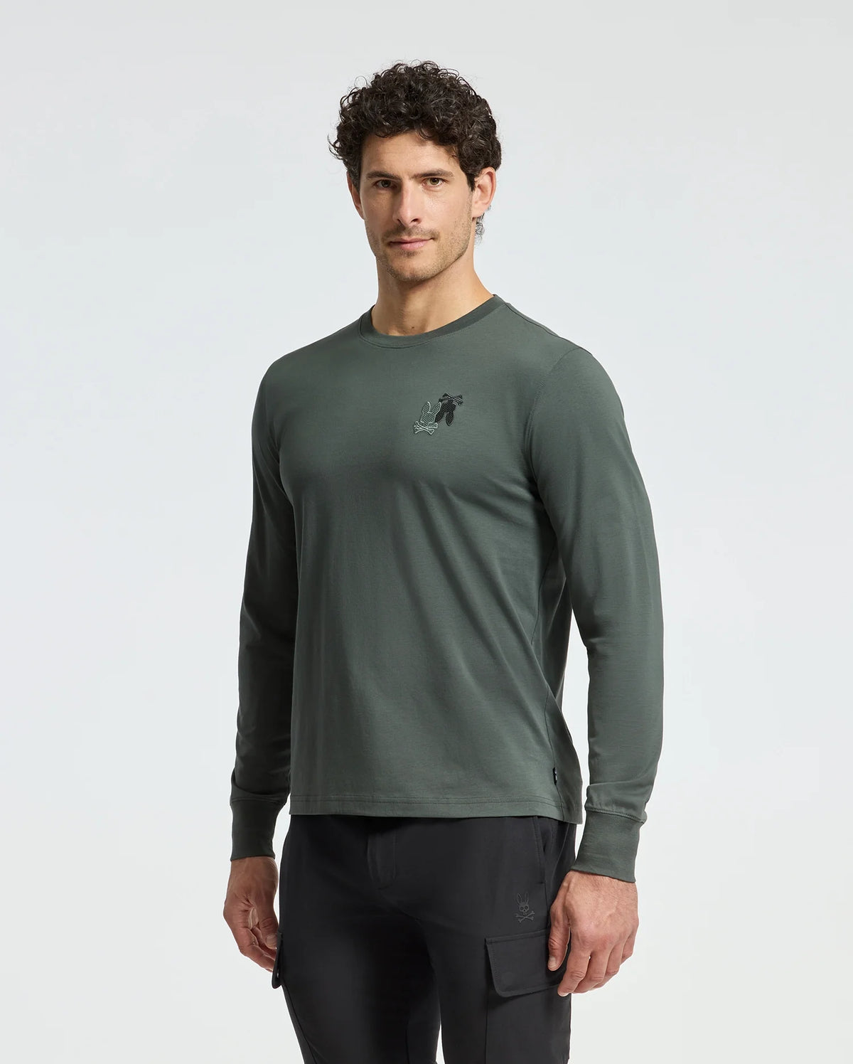 A man in a Psycho Bunny MENS PIERCE LONG SLEEVE BACK GRAPHIC TEE, dark green pima cotton with a chest graphic, and black cargo pants stands against a plain background. His curly hair complements the tee as he stands relaxed, gazing forward.