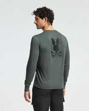 A man with curly hair wears the Psycho Bunny MENS PIERCE LONG SLEEVE BACK GRAPHIC TEE. Made of comfortable Pima cotton, it features an embroidered black bunny logo with crossed bones on the back. The dark green fabric enhances his style as he stands against a plain background, slightly to the side.