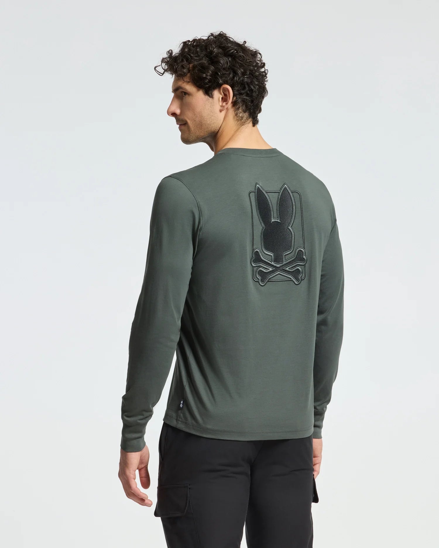 A man with curly hair wears the Psycho Bunny MENS PIERCE LONG SLEEVE BACK GRAPHIC TEE. Made of comfortable Pima cotton, it features an embroidered black bunny logo with crossed bones on the back. The dark green fabric enhances his style as he stands against a plain background, slightly to the side.